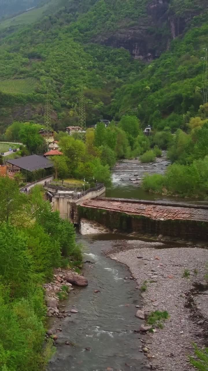 Italian village near flowing river with dam and green mountains in background, aerial vertical view