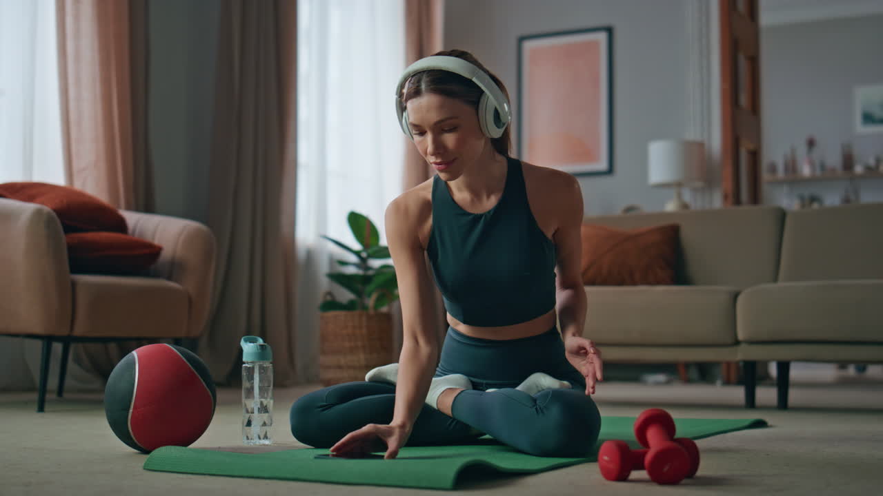 Yoga woman turning music smartphone practicing lotus position home in headphones