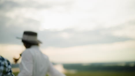 back view of a couple holding hands and running through a tall grass field. the man is in a black hat and checkered shirt, while the woman wears a white dress and hat, capturing a joyful