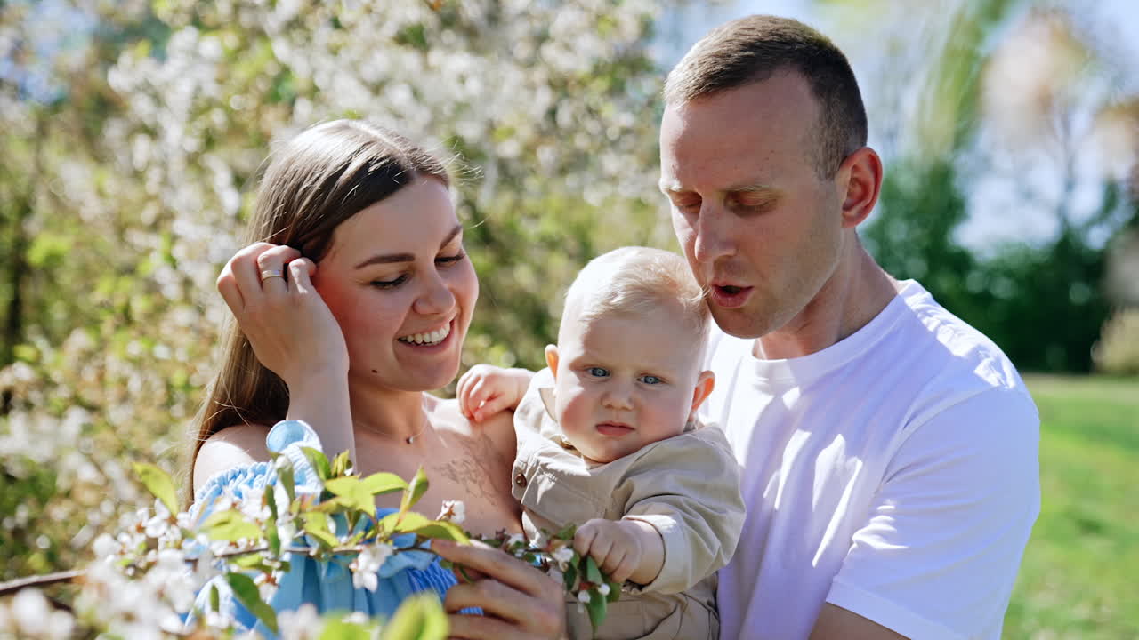 Happy parents of a little baby stand in the spring garden. Lovely child touches the tree branch.