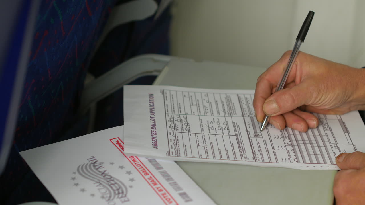 Filling out an absentee ballot application with a pen, while the "Official Election Mail" envelope rests nearby. The image highlights the process of completing a mail-in voting form during travel