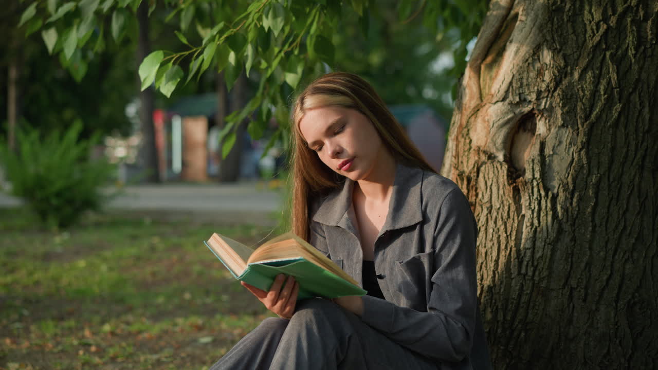 dama de ropa gris sentada al aire libre revisa un libro, mirando pensativa con la cabeza inclinada ligeramente a la derecha, el fondo presenta vegetación y un edificio borroso