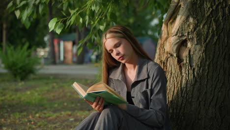 dama de ropa gris sentada al aire libre revisa un libro, mirando pensativa con la cabeza inclinada ligeramente a la derecha, el fondo presenta vegetación y un edificio borroso