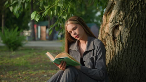 lady in grey clothing seated outdoors flips through a book, looking thoughtful with her head tilted slightly to the right, the background features greenery and a blurred building