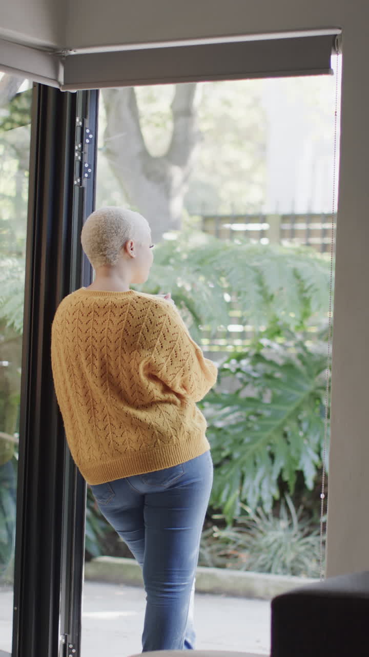 Vertical video of happy biracial woman at window drinking coffee in slow motion