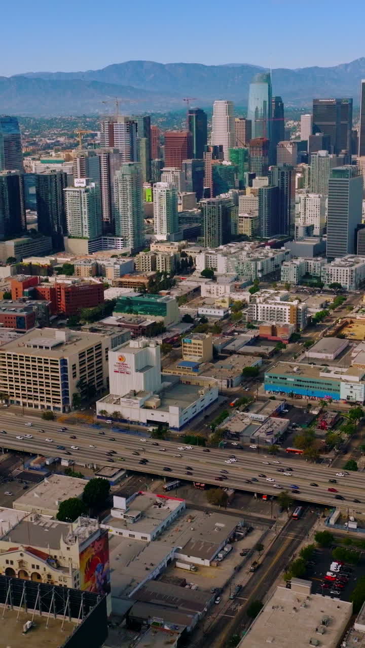Famous skyscrapers standing among the low buildings of Los Angeles, California, USA. Numerous cars moving by the freeway at daytime. Aerial view. Vertical video