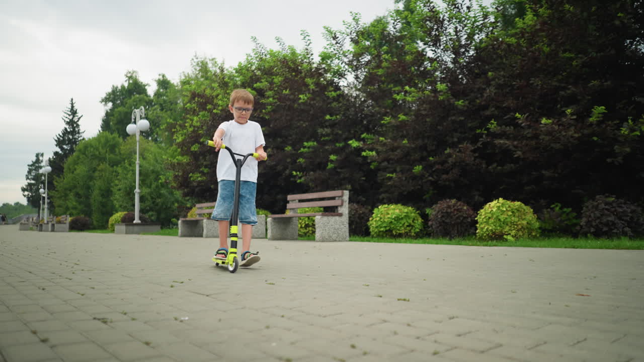 A young boy skillfully turns his scooter around on a paved path in a park, with trees and lampstand in the background