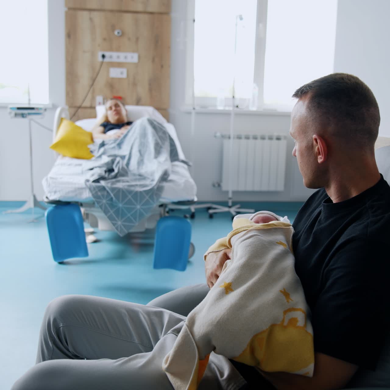 Father is holding a newborn sleeping baby in the hospital ward. Woman is resting in bed at backdrop in blur