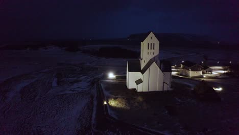 a calm nighttime aerial of a traditional icelandic church, surrounded by snowy fields and quiet rural buildings under the soft glow of winter lights.