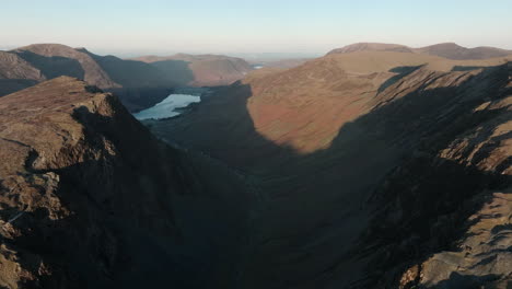 Flying-high-over-shadowed-deep-mountain-pass-with-distant-lake-and-other-mountains-visible