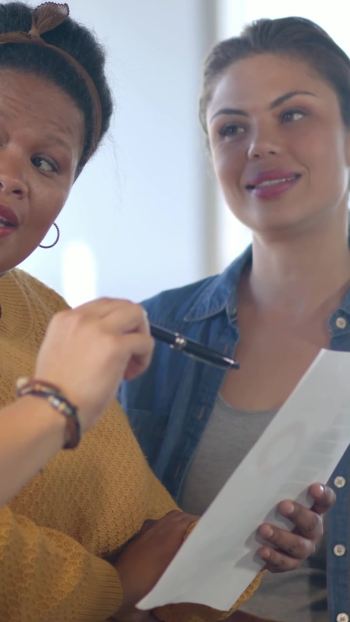Vertical video: Turning offered object, women in mustard and denim checking paper with pen in room