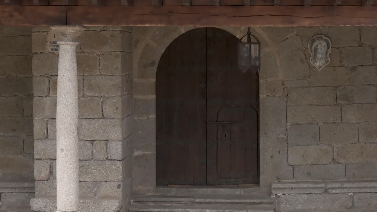 Drone shot moving slowly backward from the 15th-century Sanctuary of Nuestra Señora del Espino in Hoyos del Espino, Ávila, Spain. Granite walls, wooden door, portico with columns and sacred details