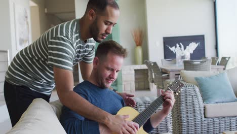 una pareja de hombres homosexuales multiétnicos sentados en el sofá tocando la guitarra juntos