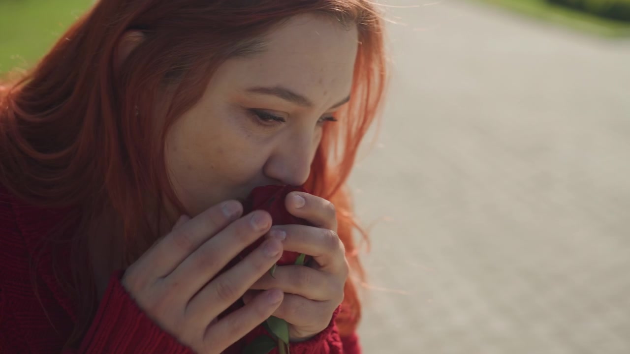 Close-up de jovem mulher ruiva usando suéter de tricô, segurando e cheirando uma rosa artificial ao ar livre. Sua expressão é contemplativa enquanto a mulher interage com a flor sob a luz suave do dia