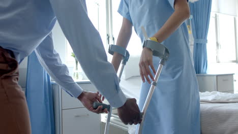 midsection of diverse male doctor giving female patient crutches in hospital ward, slow motion