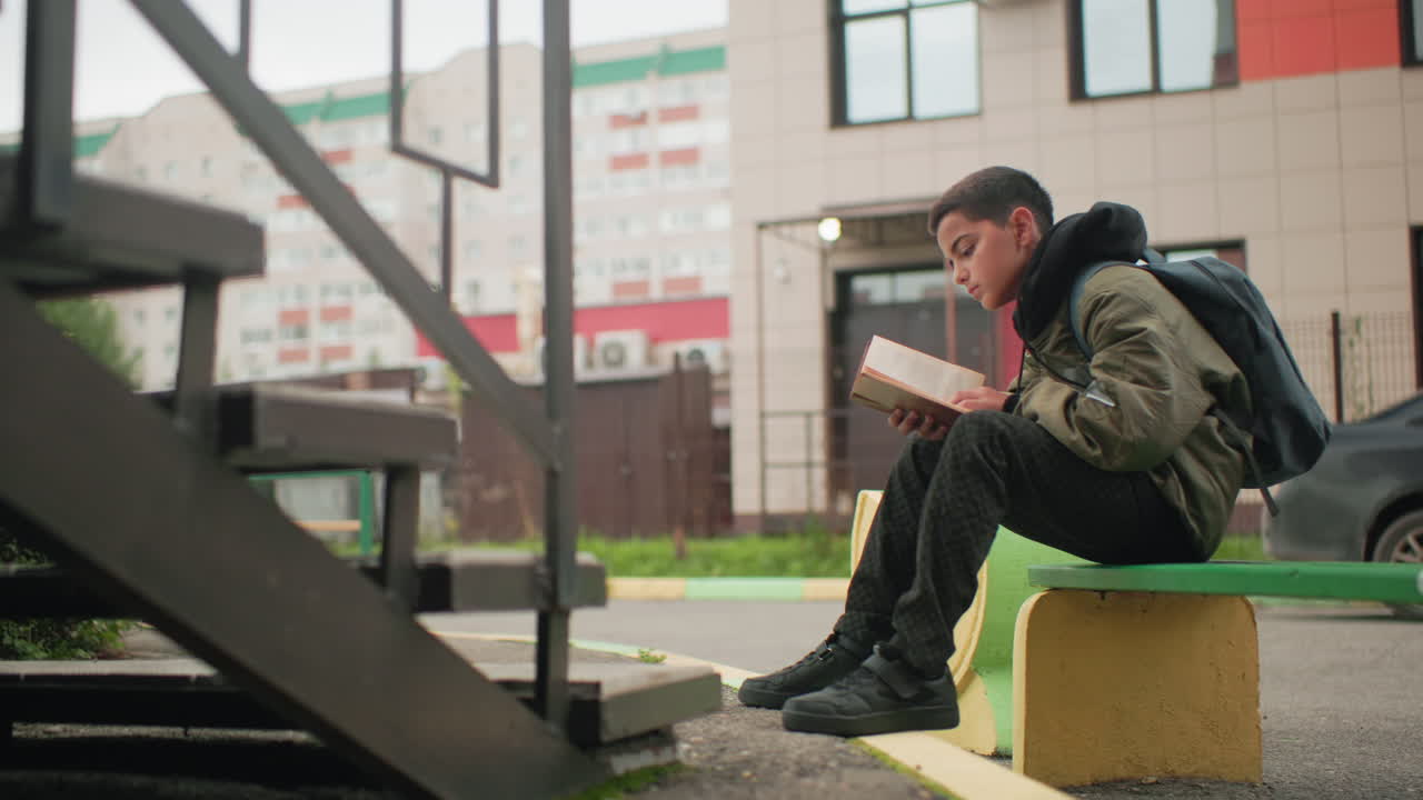 Boy seated on bench wearing green jacket and backpack focusing on study while flipping through book outdoors with blurred city building in background