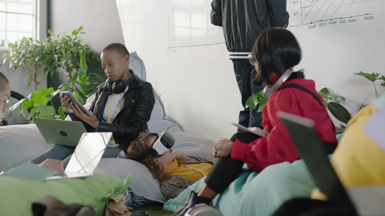 젊은 비즈니스 그룹 학생들이 태블릿 컴퓨터를 사용하는 아시아 여성 아프리카계 미국인 동료를 보여주고 바쁜 스타트업 사무실에서 창의적인 아이디어를 지원하는 조언을 공유합니다.