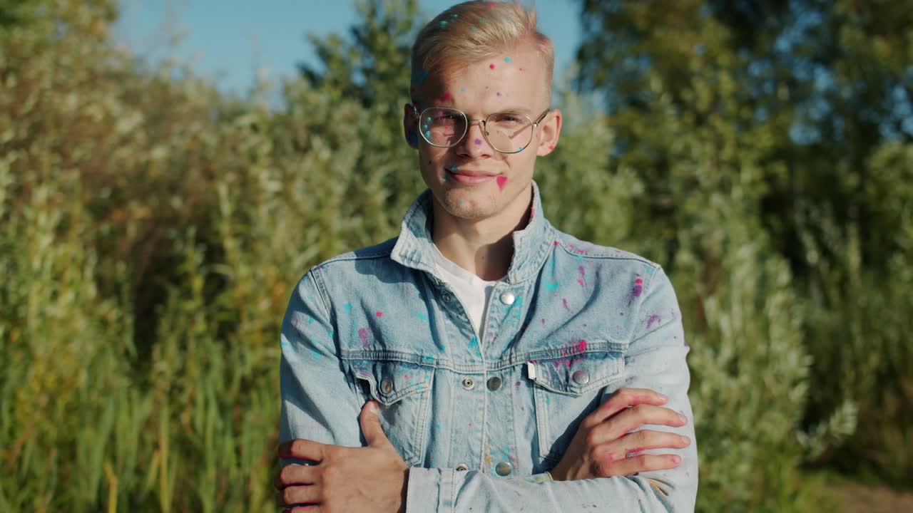 Slow motion portrait of blond man standing at Holi festival smiling and laughing when people throwing paint at him