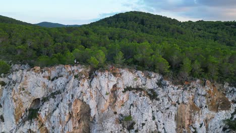 Hikers-rest-on-a-rocky-cliff-among-trees-with-a-view-of-the-green-landscape-during-sunset-in-ibiza