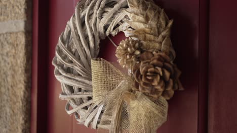 Rustic-wreath-with-burlap-bow-and-pinecones,-hanging-on-a-red-door,-exuding-cozy-charm