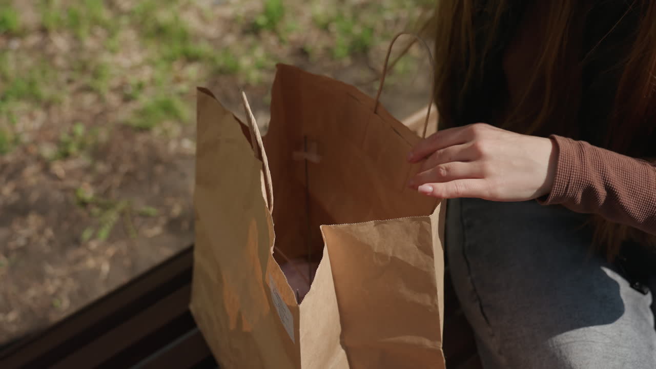 Primer plano de una persona que maneja hábilmente un hilo de colores, con las manos enhebrando lana roja junto a una bolsa de papel al aire libre; vista detallada de artesanía amateur con lana roja vibrante cerca de una bolsa de papel al aire libre