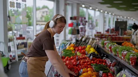 Side-view-of-a-happy-girl-wearing-wireless-headphones-in-a-brown-t-shirt-and-apron-listening-to-music-and-arranging-vegetables-on-the-counter-during-her-work-and-in-the-supermarket