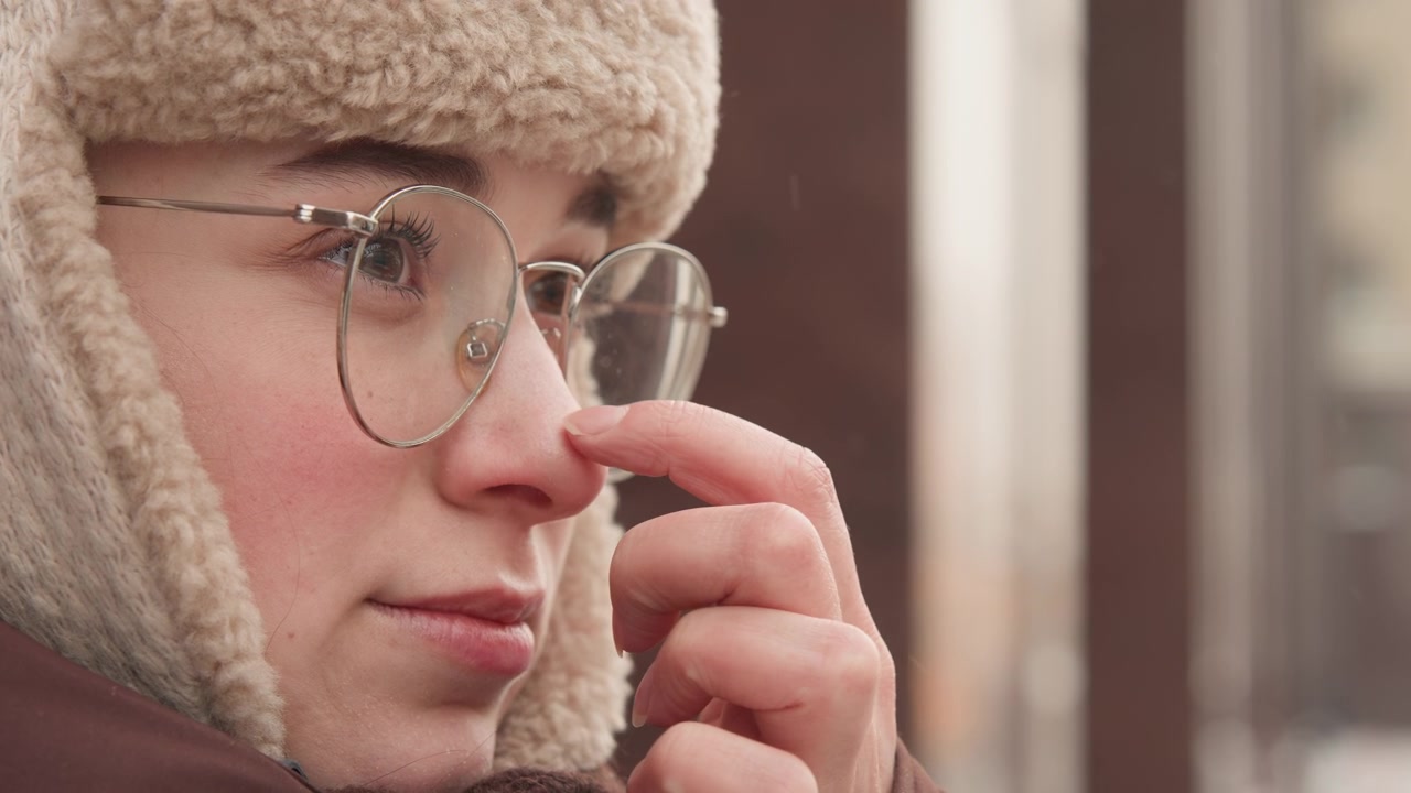 Primo piano di profilo a tre quarti mostra una giovane persona che gira delicatamente la testa e usa la punta delle dita per aggiustare occhiali rotondi in metallo sul naso mentre indossa un cappello di pile testurizzato con paraorecchie all'aperto.