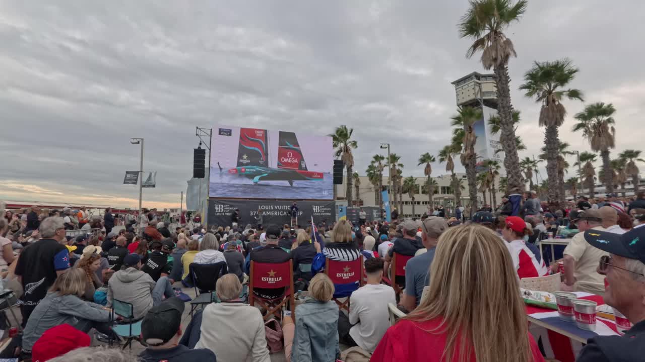 Crowd outside watching the 37th America´s cup on a giant screen. Celebrations. On the screen you can see a sailboat at high speed