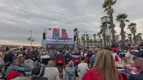crowd outside watching the 37th america´s cup on a giant screen. celebrations. on the screen you can see a sailboat at high speed