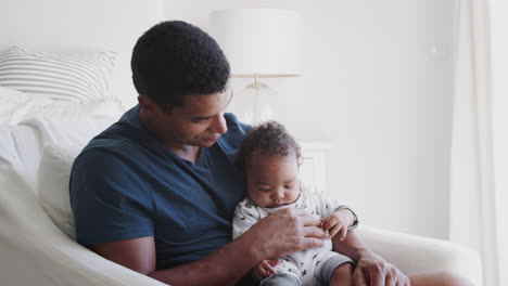 millennial african american dad sitting in an armchair holding his baby boy, close up, side view