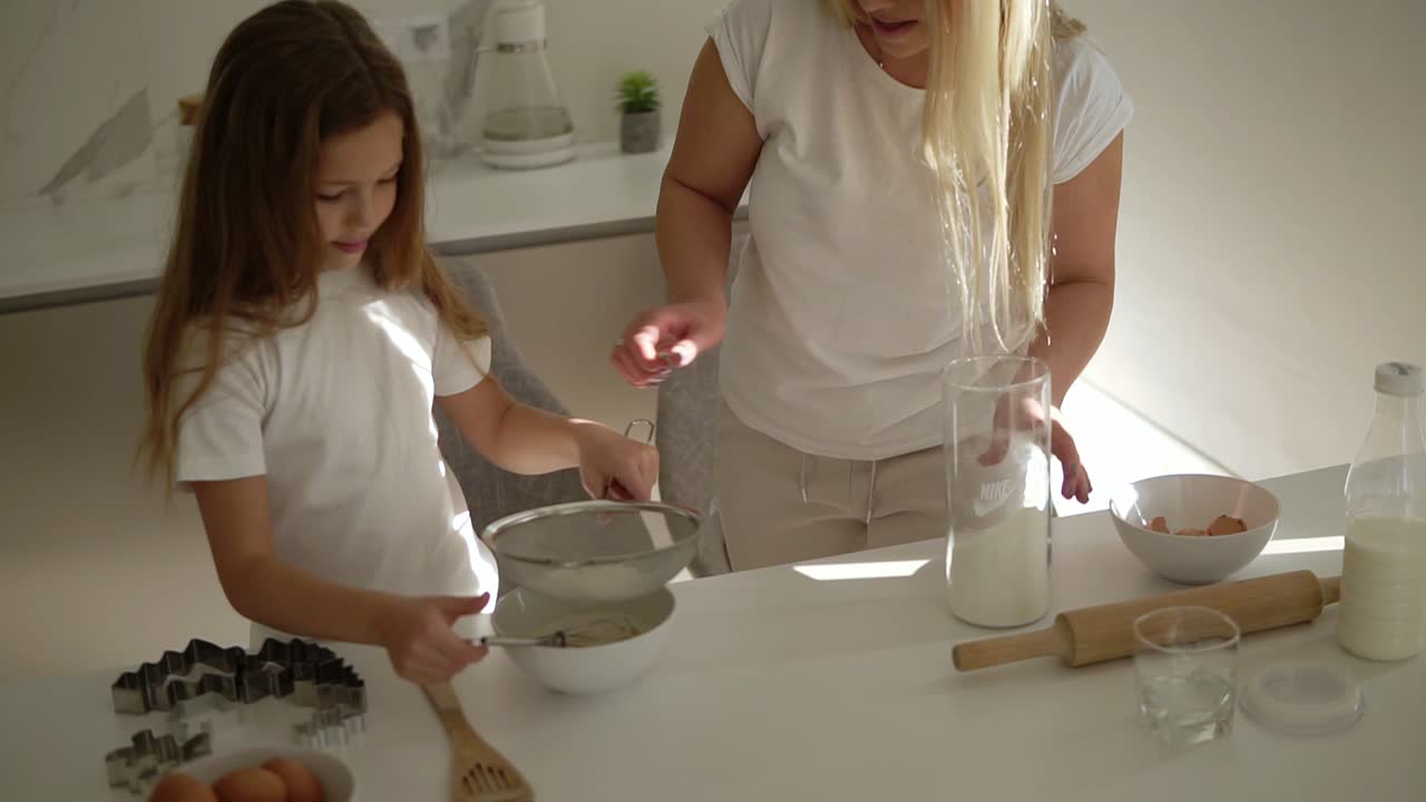 Mother helping and teaching her cute daughter in modern cozy white kitchen to cook cake or pancakes. Mother adding flour through a sieve and girl mixing ingredients. Happy family. Relationship mom and daughter