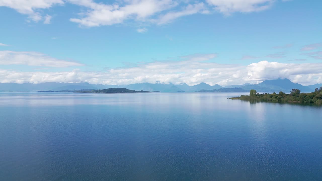 チリ、ランコ湖。島々と雪をかぶったアンデス山脈の峰々がある鏡のような湖上空を降下するショット