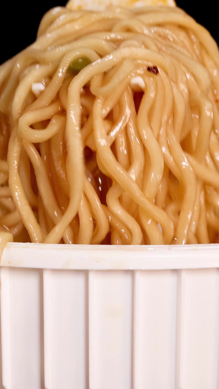 Una mano usa le bacchette per sollevare noodles istantanei cotti da un contenitore a tazza bianco su uno sfondo nero, sotto un'illuminazione brillante e uniforme, con una prospettiva ravvicinata.