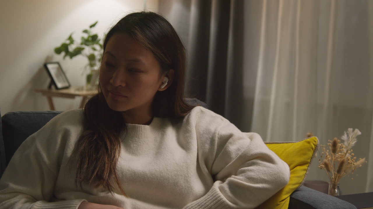 소파에서 집에서 저녁을 보내는 여성, 무선 이어폰을 착용하고 음악이나 캐스트를 스트리밍합니다.