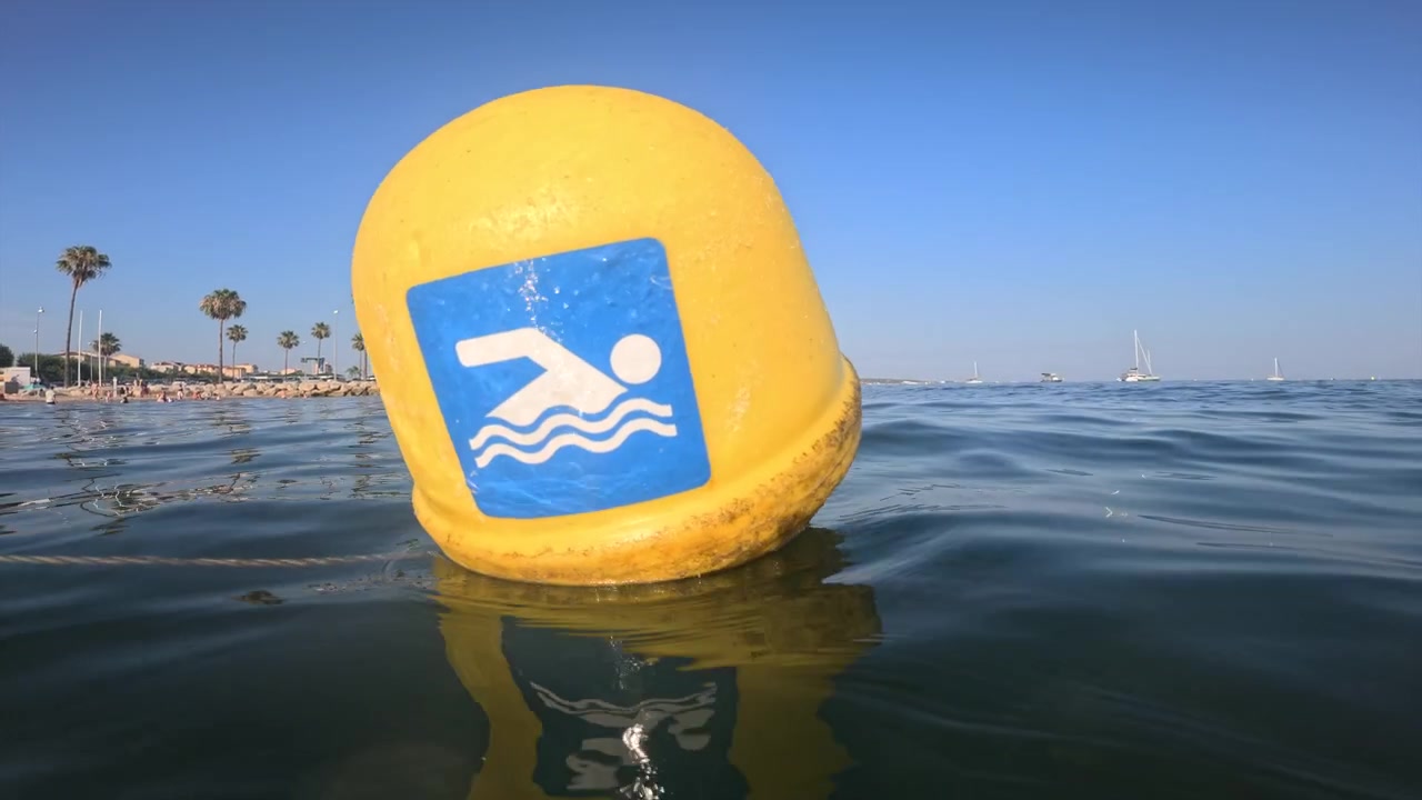 Pelampung kuning menandai area berenang yang ditentukan di laut dengan pantai berpasir dan pohon palem di latar belakang selama musim panas