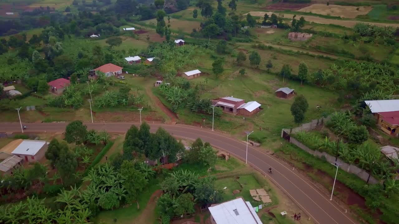 Drone tracking shot of a motorcycle riding along a curved road lined with banana plants and houses near Mount Elgon in Uganda, with fields, trees, and rural buildings forming a lush green landscape
