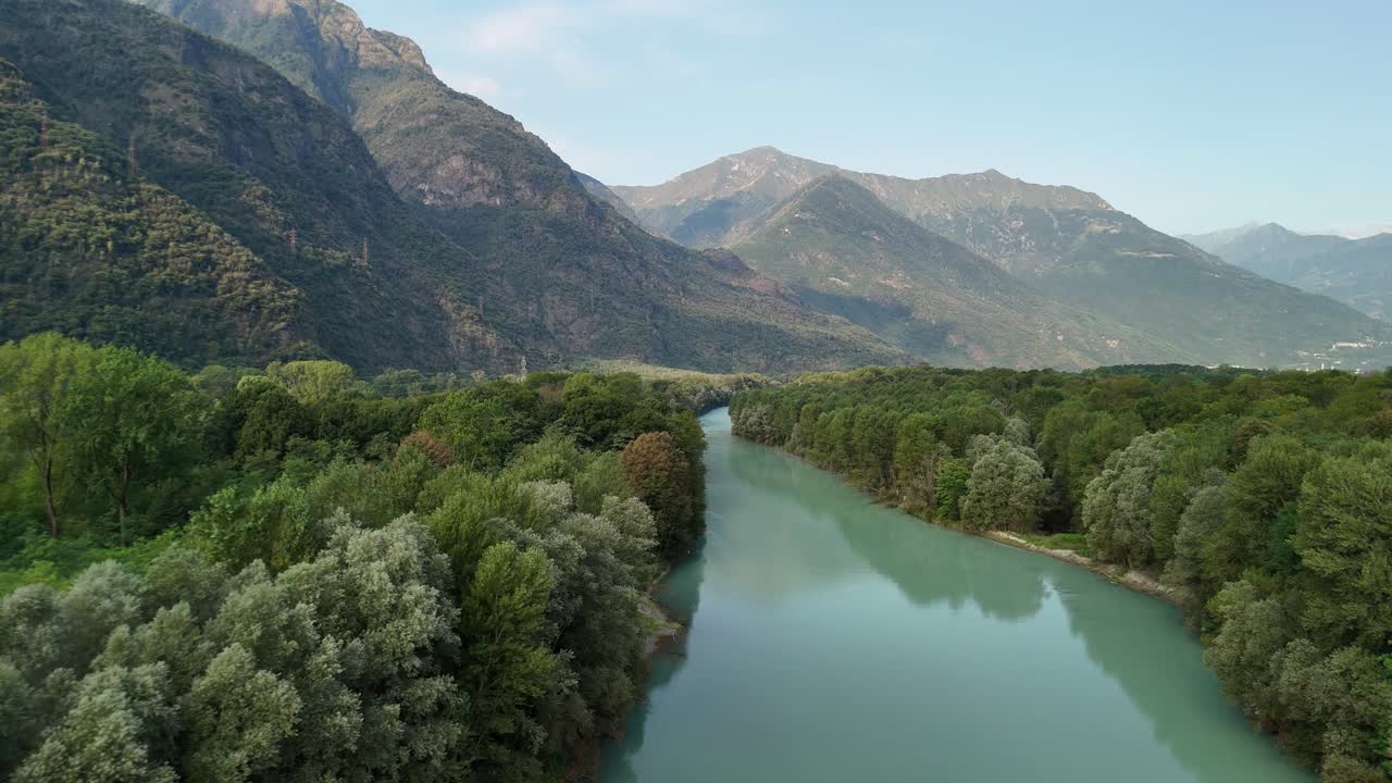 A drone pans down over the Toce River, nestled between trees, with majestic mountains and a clear sky in the background, creating a breathtaking view of nature in Ansola, Italy