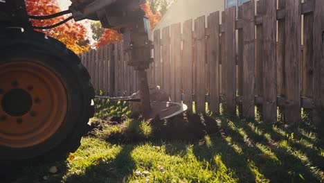 auger digging a hole to plant a tree on a sunny day with colorful trees and a wooden fence in the background