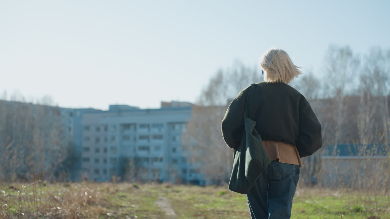 Woman with blonde hair in dark coat walking outdoors with back turned, sunlight shining, urban buildings in background, casual lifestyle moment capturing independence, solitude, and thoughtful atmosphere