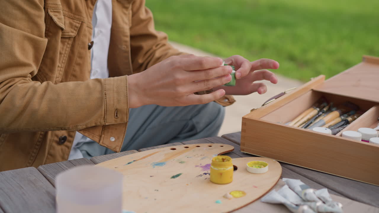 Scena all'aperto con palette e scatola di colori che mostra mani che selezionano pennelli e spremono tubetti, mescolando giallo e verde su una palette di legno accanto a una scatola aperta di materiali su un tavolo da picnic in un parco, in un'atmosfera rilassata e creativa