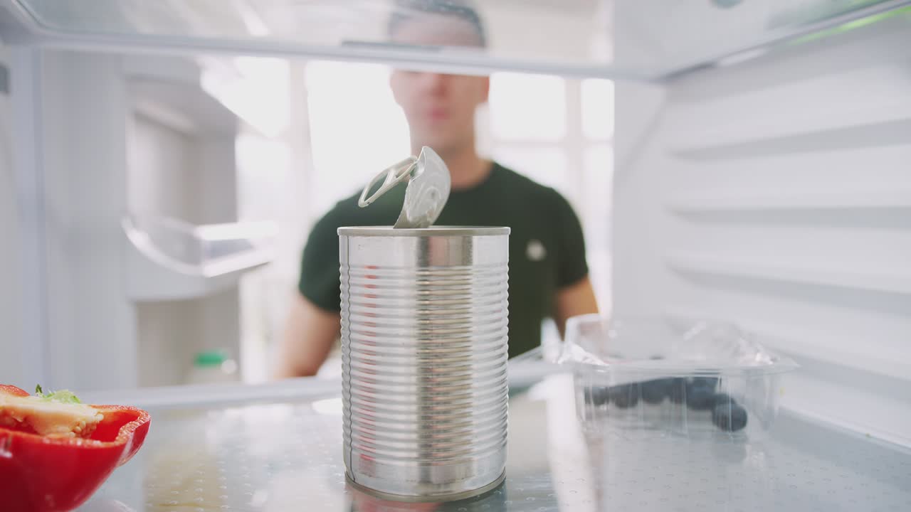 joven poniendo la lata abierta de nuevo en el estante en el refrigerador vacío