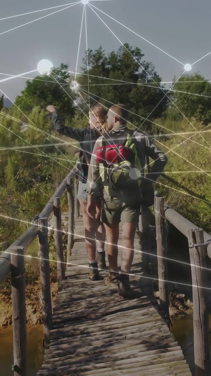 Video verticale: Donna che indica avanti, uomo che scansiona escursionisti che camminano su un ponte, con nodi tecnologici sovrapposti alla scena.