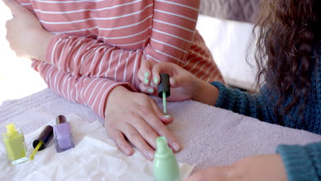 Happy-biracial-daughter-sitting-and-painting-mother's-nails-in-sunny-room