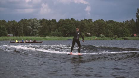 Mann-Auf-Folie-Surfen-In-Welle-Mit-Ruderer-Im-Hintergrund