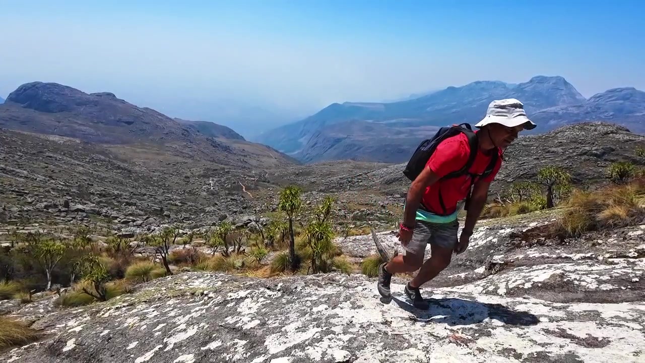 Un hombre con mochila trepa por una roca inclinada en el monte Mulanje, Malaui, con un amplio valle montañoso y crestas escarpadas detrás de él
