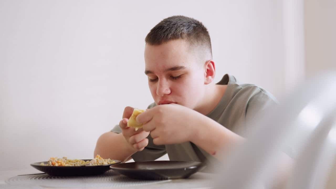 Young boy in green shirt eagerly eats folded pancake with cream while interacting with person off screen, sitting at dining table with other food on plate, enjoying mealtime in sunlit home setting