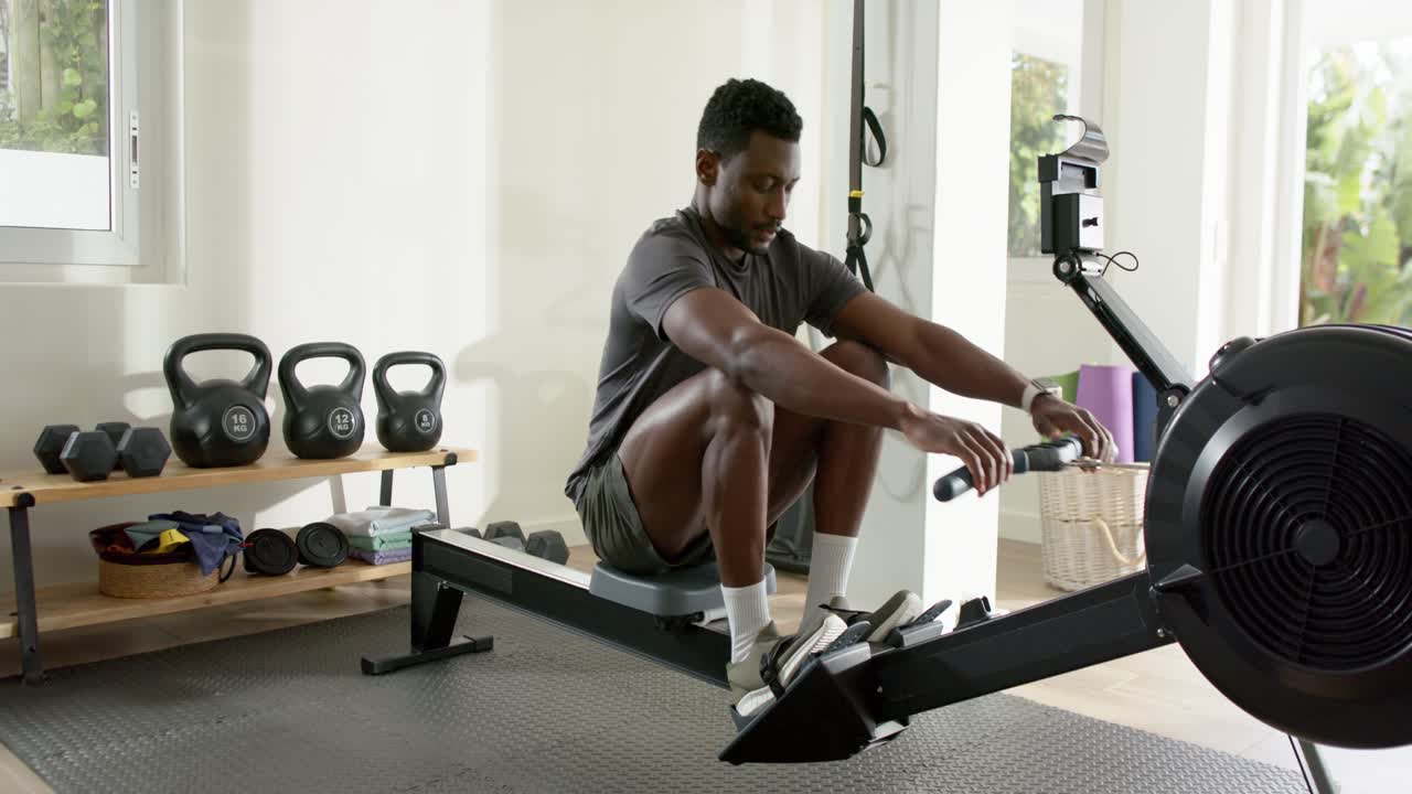 Mid-adult African American man pressing heels, pulling handle on rower in home gym for strength
