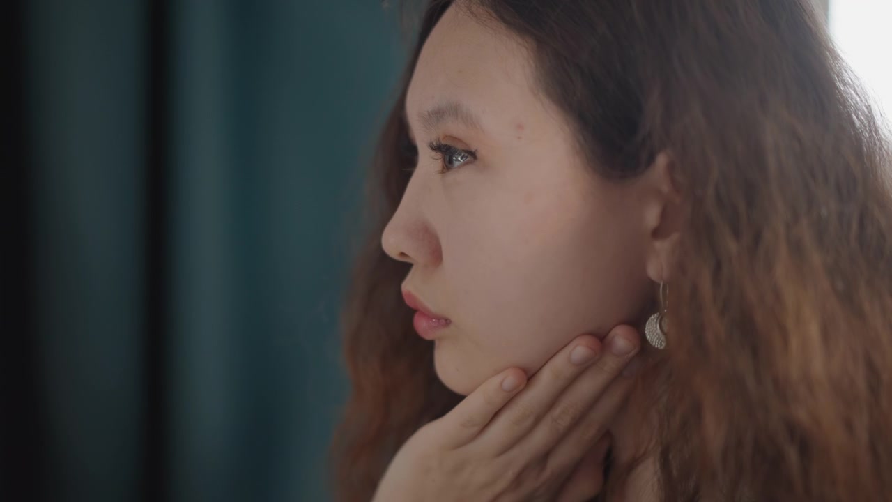 Close-up side profile shows young woman with closed eyes, long wavy brown hair, dangling earring, and hand at neck, set against soft indoor light, teal tones, shallow depth, quiet mood