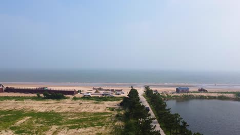 Vista-Aérea-De-Pedestal-Ascendente-De-La-Playa-De-Arena-De-Nanhai-Con-Estanque-En-El-Lado-Derecho-De-La-Carretera-Y-Césped-En-El-Lado-Izquierdo