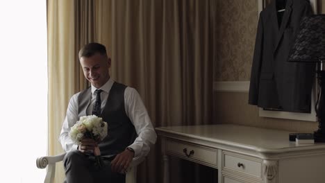 handsome groom man with wedding bouquet in his hands at home near window preparing to go to bride